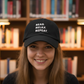 Person wearing a black cap with 'READ. RELAX. REPEAT.' text in a library setting