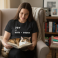 woman reading a book with a cat on her lap wearing a Joy = Cats + Books black t-shirt.